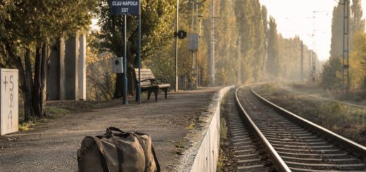 An early autumn morning on a quiet suburban railway platform at the edge of a Romanian university city. A single canvas duffel bag stands on the worn concrete edge of the platform, low warm sunlight raking along the polished rails, thin mist drifting over a line of poplars behind, an empty bench further down the platform. Shallow depth of field on the bag, the tracks curving gently away into the distance, muted green, amber and grey tones. Documentary photography, natural morning light. Use balanced, natural contrast with a realistic tonal range. Preserve fine detail throughout the image, with smooth highlight roll-off, detailed shadows, balanced exposure, and natural, accurate colors.