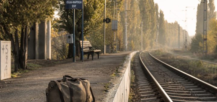 An early autumn morning on a quiet suburban railway platform at the edge of a Romanian university city. A single canvas duffel bag stands on the worn concrete edge of the platform, low warm sunlight raking along the polished rails, thin mist drifting over a line of poplars behind, an empty bench further down the platform. Shallow depth of field on the bag, the tracks curving gently away into the distance, muted green, amber and grey tones. Documentary photography, natural morning light. Use balanced, natural contrast with a realistic tonal range. Preserve fine detail throughout the image, with smooth highlight roll-off, detailed shadows, balanced exposure, and natural, accurate colors.