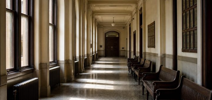 A long, quiet administrative corridor inside an old university building, photographed straight down its length in mid morning. Tall arched windows on the left throw wide bands of daylight across a worn terrazzo floor. A row of empty dark wooden benches lines the right-hand wall beneath a high ceiling with simple plaster mouldings, and a closed panelled door sits at the far end in soft focus. Still, patient atmosphere, dust drifting in the light. Architectural photography, natural available light, muted cream, oak brown and cool grey tones. Use balanced, natural contrast with a realistic tonal range. Preserve fine detail throughout the image, with smooth highlight roll-off, detailed shadows, balanced exposure, and natural, accurate colors.