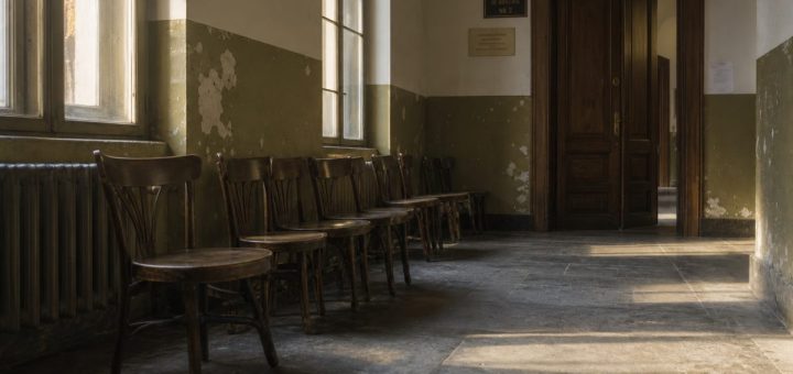 Late afternoon light in the empty waiting corridor of an old Romanian courthouse. Tall windows with simple metal frames cast long warm bands across a worn grey stone floor. A row of dark wooden chairs stands against a wall painted in faded olive green, the paint chipped at the corners. A heavy oak door with a dull brass handle is slightly ajar at the far end. Air is still, faint dust hangs in the light. Quiet, unstaged, documentary photography, shallow depth of field, muted palette of ochre, olive and grey. Use balanced, natural contrast with a realistic tonal range. Preserve fine detail throughout the image, with smooth highlight roll-off, detailed shadows, balanced exposure, and natural, accurate colors.