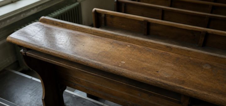 A single empty wooden lecture-hall bench seen from above at a slight angle, worn varnish catching soft window light from the left, faint scuff marks on the surface, rows of identical benches falling away into shallow depth of field behind. Restrained palette of honey oak, cool grey and muted olive. Documentary interior photography, natural indirect daylight, understated and serious. Use balanced, natural contrast with a realistic tonal range. Preserve fine detail throughout the image, with smooth highlight roll-off, detailed shadows, balanced exposure, and natural, accurate colors.