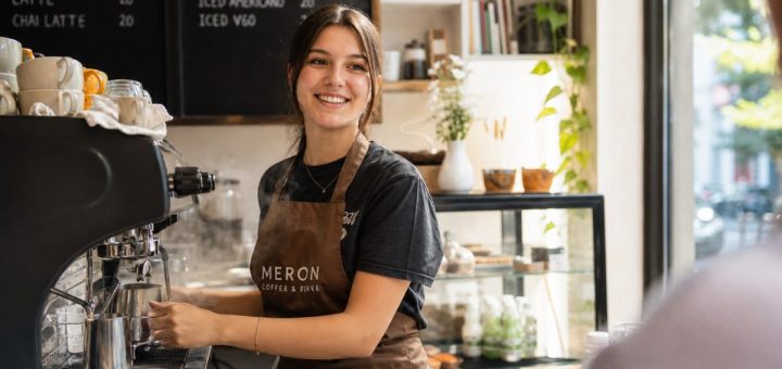 A cheerful young Romanian student wearing a barista apron working behind the counter of a bright neighbourhood café, steam rising from an espresso machine, smiling toward a customer just out of frame, warm daylight pouring through the front window. Candid reportage photography, natural skin tones. Use balanced, natural contrast with a realistic tonal range. Preserve fine detail throughout the image, with smooth highlight roll-off, detailed shadows, balanced exposure, and natural, accurate colors.