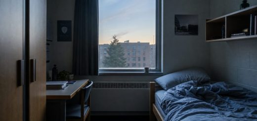 An empty student dormitory room at dawn, soft blue-grey light coming through a half-drawn curtain, an unmade bed with rumpled sheets, a ceramic cup left on the windowsill, still and quiet atmosphere, interior architectural photography. Use balanced, natural contrast with a realistic tonal range. Preserve fine detail throughout the image, with smooth highlight roll-off, detailed shadows, balanced exposure, and natural, accurate colors.