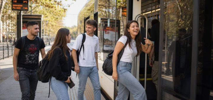 A group of cheerful Romanian university students in casual clothing stepping onto a modern low-floor city tram at a bright urban stop, warm morning light on the pavement, a European city street lined with plane trees behind them, one student laughing while holding the handrail, backpacks over their shoulders. Candid reportage photography, shallow depth of field. Use balanced, natural contrast with a realistic tonal range. Preserve fine detail throughout the image, with smooth highlight roll-off, detailed shadows, balanced exposure, and natural, accurate colors.