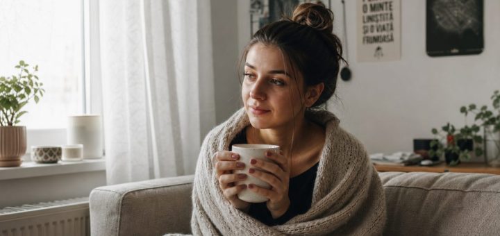 Candid reportage photograph of a young Romanian student in her early twenties wrapped in a soft knitted blanket on a sofa in a small tidy sunlit apartment, holding a warm ceramic mug with both hands, tired but calm and peaceful expression, a large window on the left with sheer curtains diffusing gentle daylight, a few potted plants on the windowsill, shallow depth of field. Use balanced, natural contrast with a realistic tonal range. Preserve fine detail throughout the image, with smooth highlight roll-off, detailed shadows, balanced exposure, and natural, accurate colors.