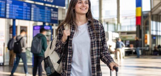 Candid reportage photograph of a smiling young Romanian woman student in her early twenties standing in a bright modern railway station hall with a small wheeled suitcase beside her, canvas backpack on one shoulder, looking ahead with excitement, large glass windows and travellers softly blurred in the background, clean morning daylight, shallow depth of field. Use balanced, natural contrast with a realistic tonal range. Preserve fine detail throughout the image, with smooth highlight roll-off, detailed shadows, balanced exposure, and natural, accurate colors.