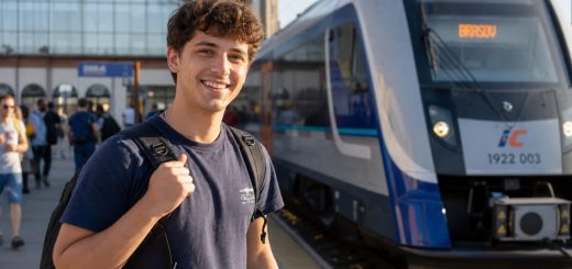 Candid reportage photograph of a happy young Romanian man student in his early twenties standing on a sunlit open railway station quay with a backpack over one shoulder, smiling as a modern train arrives beside him, other travellers and station architecture softly blurred in the background, warm golden light, shallow depth of field. Use balanced, natural contrast with a realistic tonal range. Preserve fine detail throughout the image, with smooth highlight roll-off, detailed shadows, balanced exposure, and natural, accurate colors.