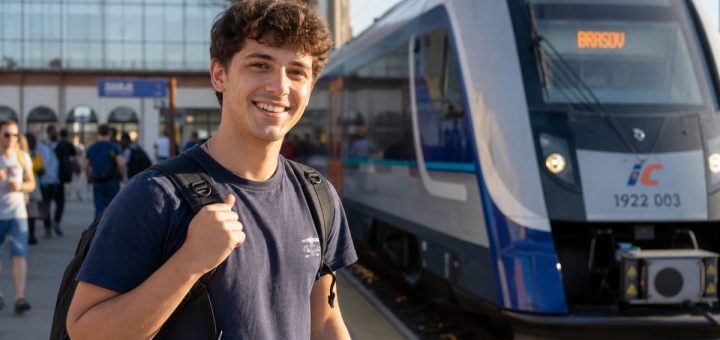 Candid reportage photograph of a happy young Romanian man student in his early twenties standing on a sunlit open railway station quay with a backpack over one shoulder, smiling as a modern train arrives beside him, other travellers and station architecture softly blurred in the background, warm golden light, shallow depth of field. Use balanced, natural contrast with a realistic tonal range. Preserve fine detail throughout the image, with smooth highlight roll-off, detailed shadows, balanced exposure, and natural, accurate colors.